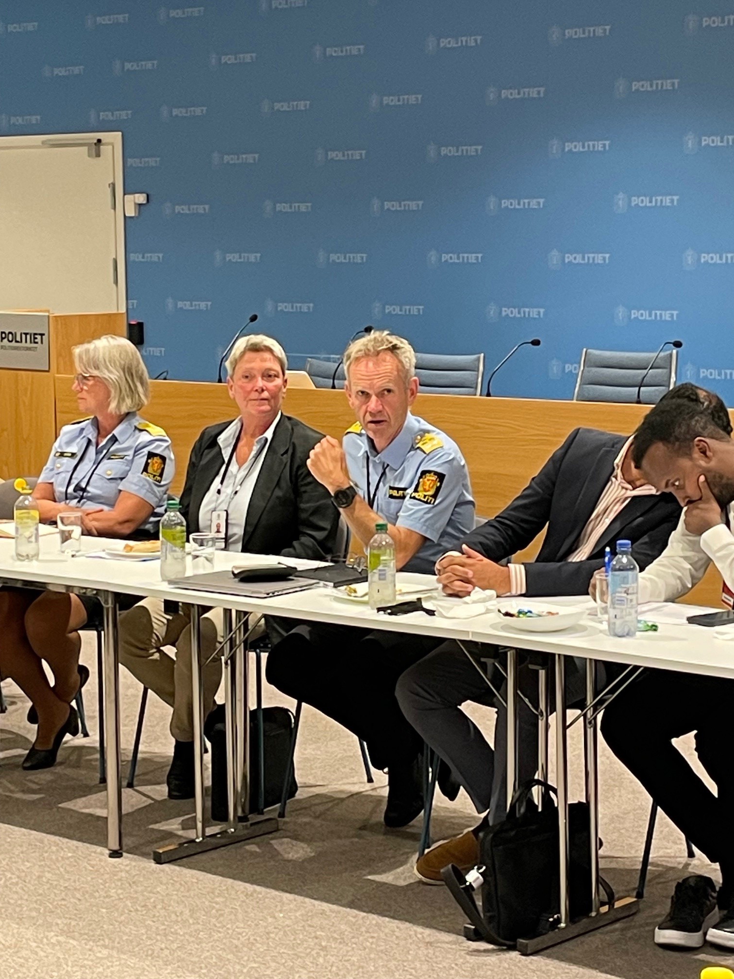 To i politiuniform og to i dress på et bor. Mann i politiuniform snakker.